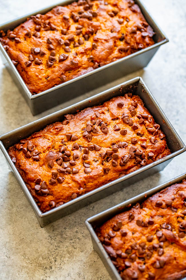 Three rectangular loaf pans filled with chocolate chip banana bread are pictured on a light-colored surface. The golden-brown loaves have chocolate chips scattered on top.