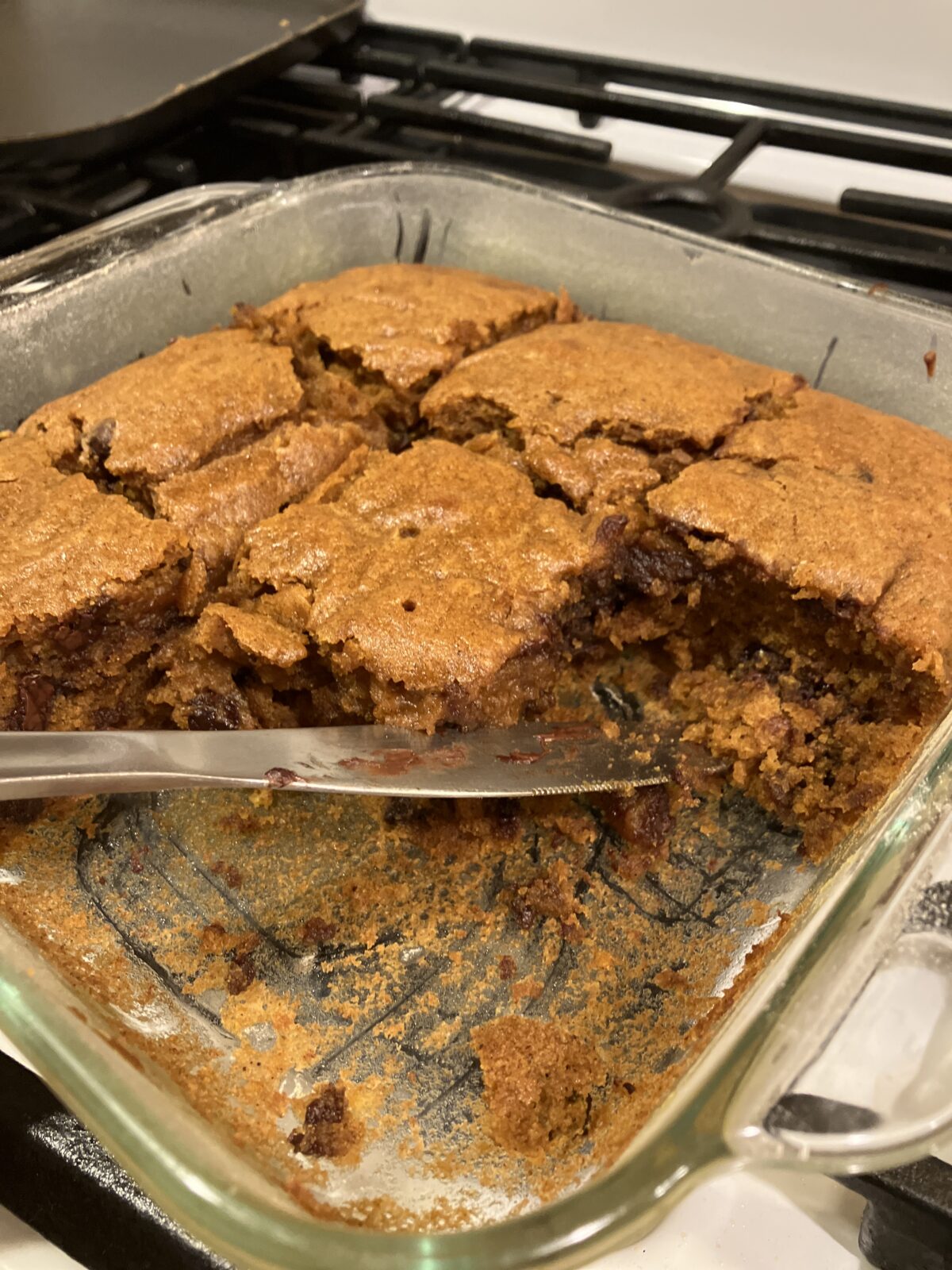 A glass dish with partially cut brown baked dessert squares and a metal knife, resting on a stove.