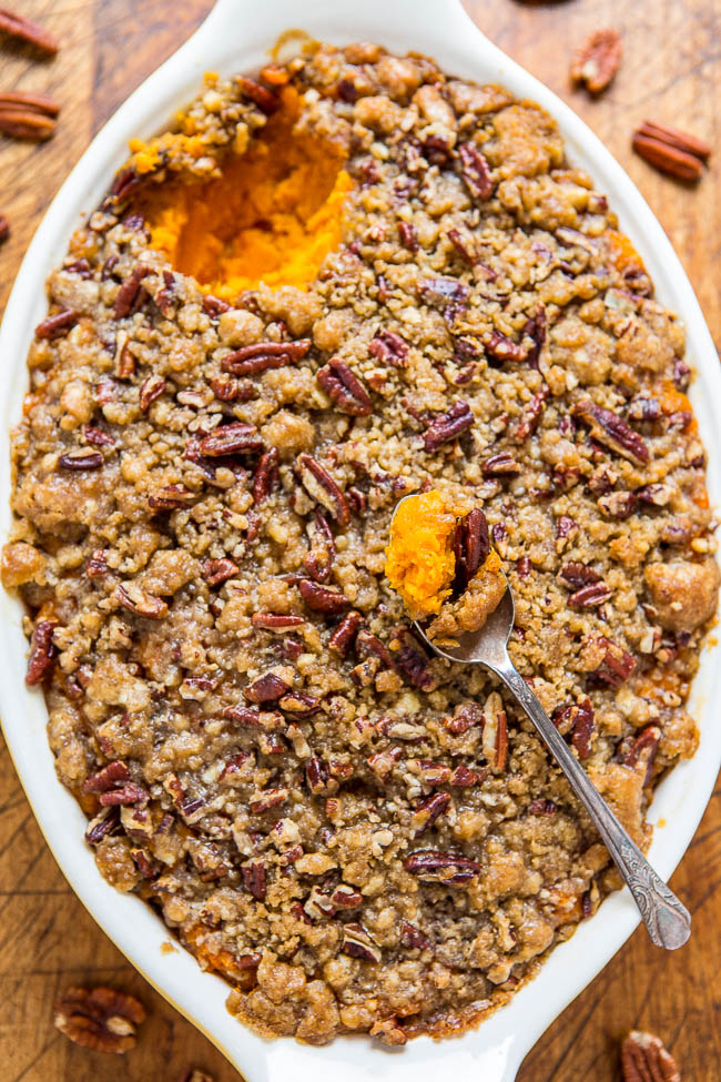 A white baking dish filled with a sweet potato casserole topped with a crumbly pecan streusel. A spoon with a scoop of the casserole rests in the dish, revealing the vibrant orange sweet potatoes.