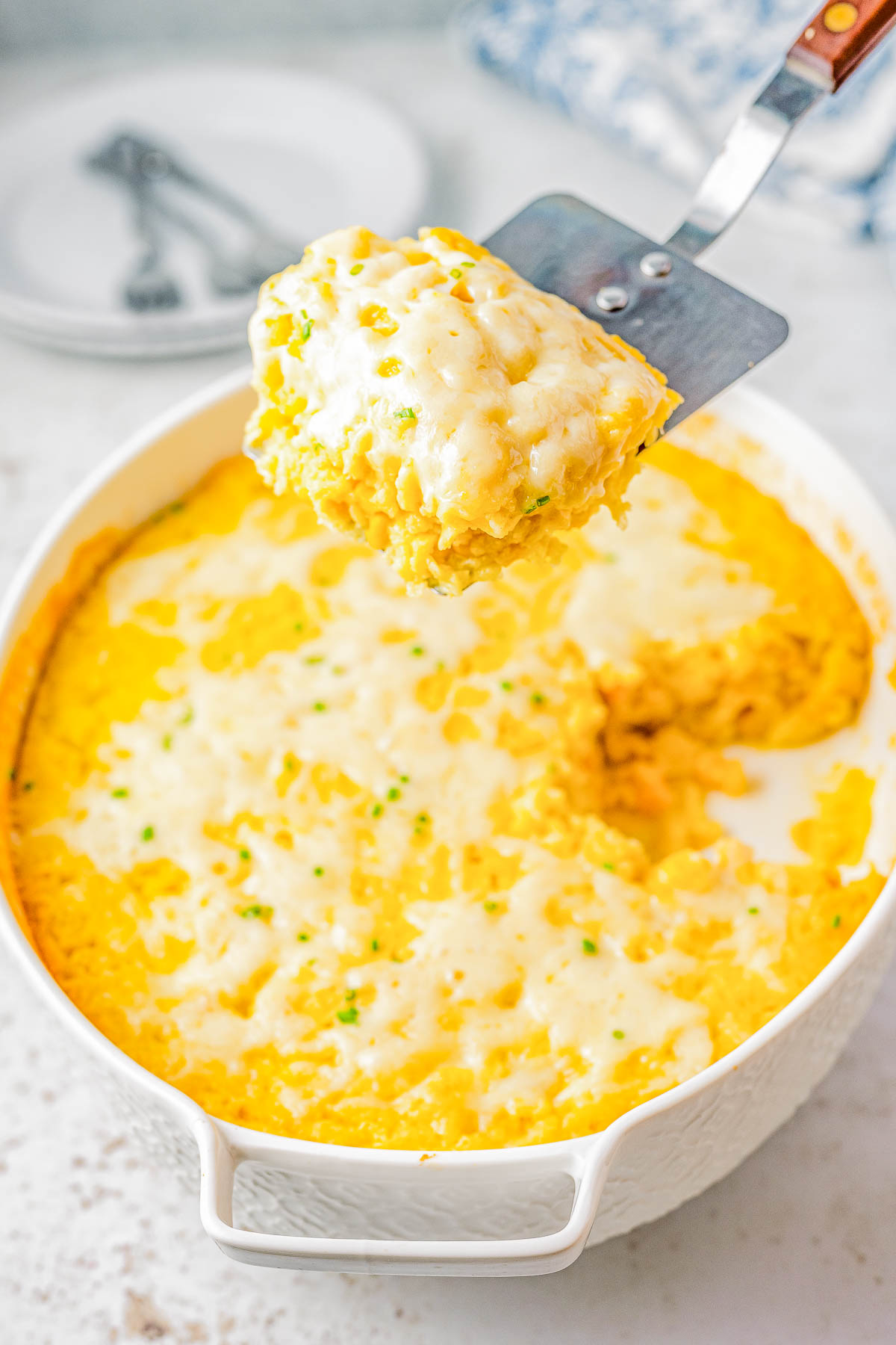 A spatula holds a piece of baked macaroni and cheese above a white casserole dish filled with the cheesy dish.