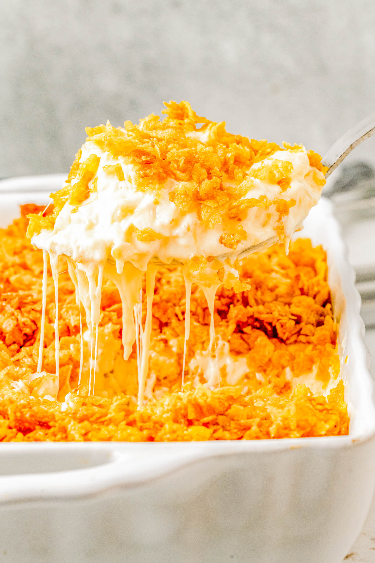 A serving spoon lifting a portion of a casserole with a golden, crispy topping and melted cheese from a white baking dish.