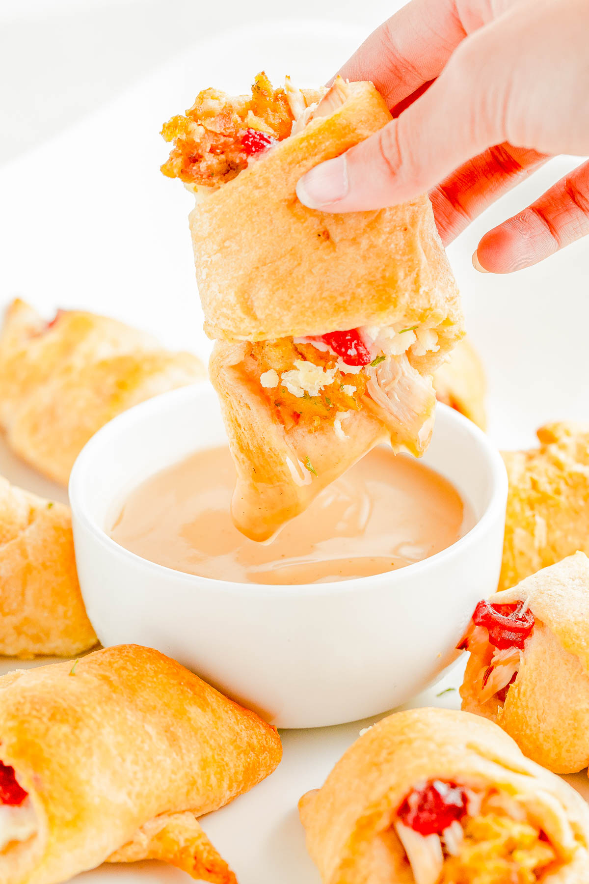 A hand dips a filled crescent roll into a bowl of dipping sauce, with other crescent rolls arranged around.