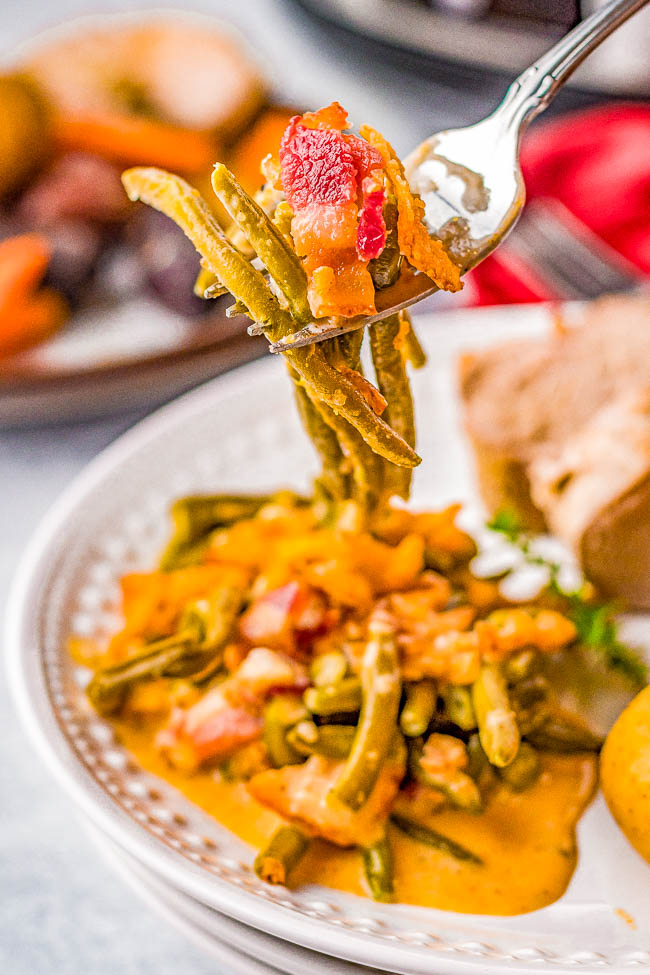 A fork holds green beans and a red bell pepper slice over a plate of cooked green beans in sauce, with bread and vegetables in the background.