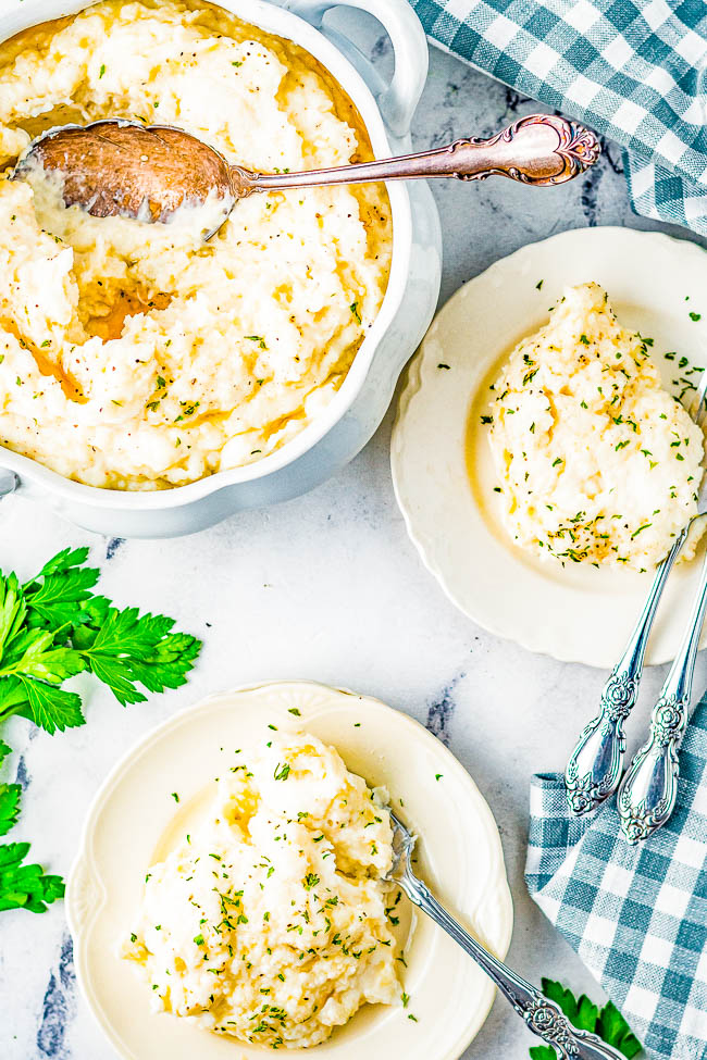 A bowl of mashed potatoes with a serving spoon, accompanied by two plates each containing a portion of mashed potatoes, garnished with herbs. A checkered cloth and fresh parsley are also visible.