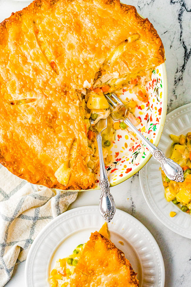 A freshly baked pot pie with a golden crust is placed on a floral-patterned dish. Two slices are served on white plates, one fork resting in the dish and another on a plate. A white cloth lies nearby.