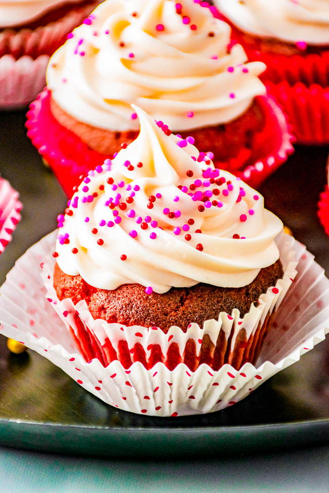 A red velvet cupcake with white frosting and pink sprinkles in a paper liner.