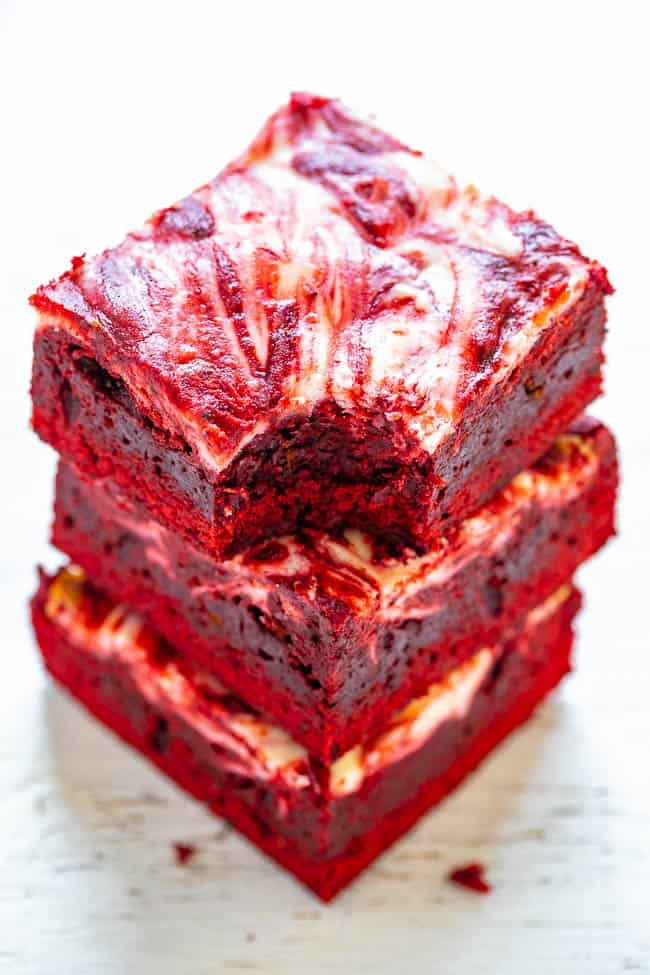 Three stacked red velvet brownies with a cream cheese swirl on top, set against a plain background.