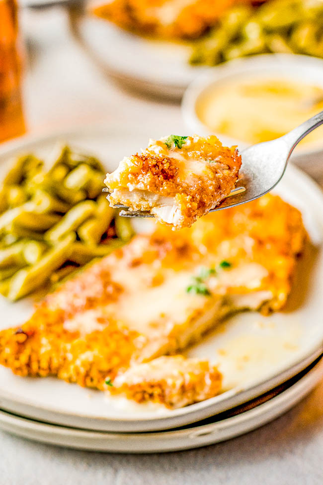 A fork holds a bite of breaded chicken over a plate with more chicken and a side of green beans.