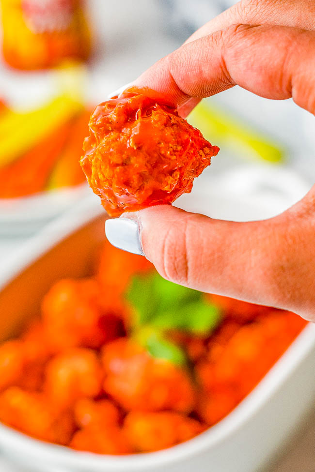 Close-up of a hand holding a buffalo chicken meatball with a dish of more meatballs and sliced carrots in the background.