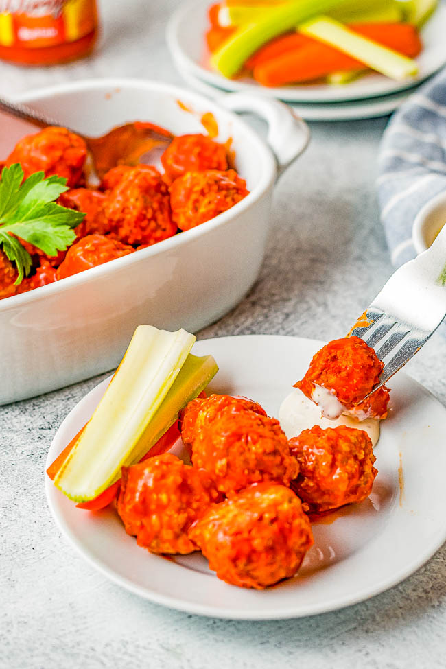 Buffalo chicken meatballs on a plate with celery sticks. A fork holds a meatball dipped in white sauce. A dish with more meatballs and a plate with carrots and celery are in the background.