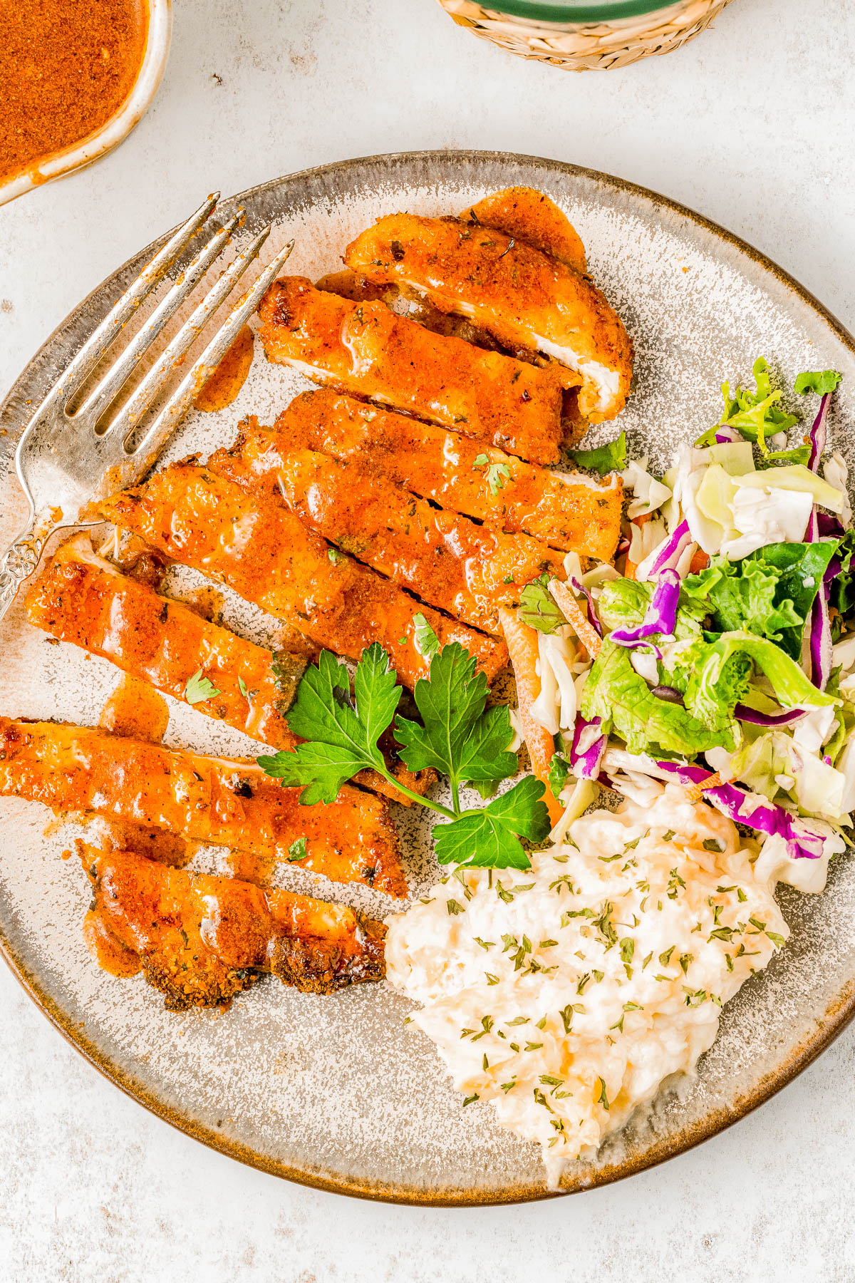 A plate with sliced grilled chicken topped with sauce, a side of coleslaw, and a mixed greens salad, garnished with parsley. A fork lies on the plate's edge.