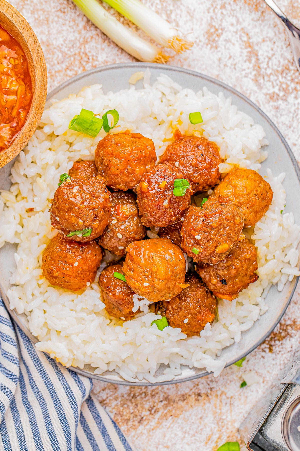 A plate of white rice topped with glazed meatballs, garnished with chopped green onions. A striped napkin and a bowl of sauce are nearby.