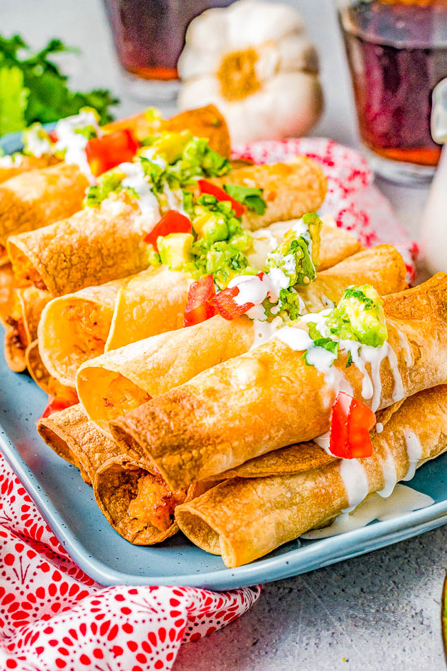 A plate of crispy taquitos topped with guacamole, diced tomatoes, and drizzled with white sauce. Garlic bulb and drinks in the background.