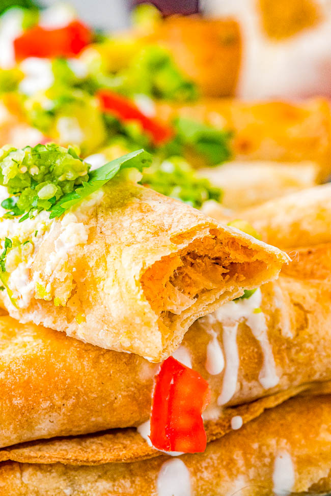 Close-up of a stack of rolled tacos topped with guacamole, diced tomatoes, and sour cream, with one taco partially bitten to reveal a meat filling.