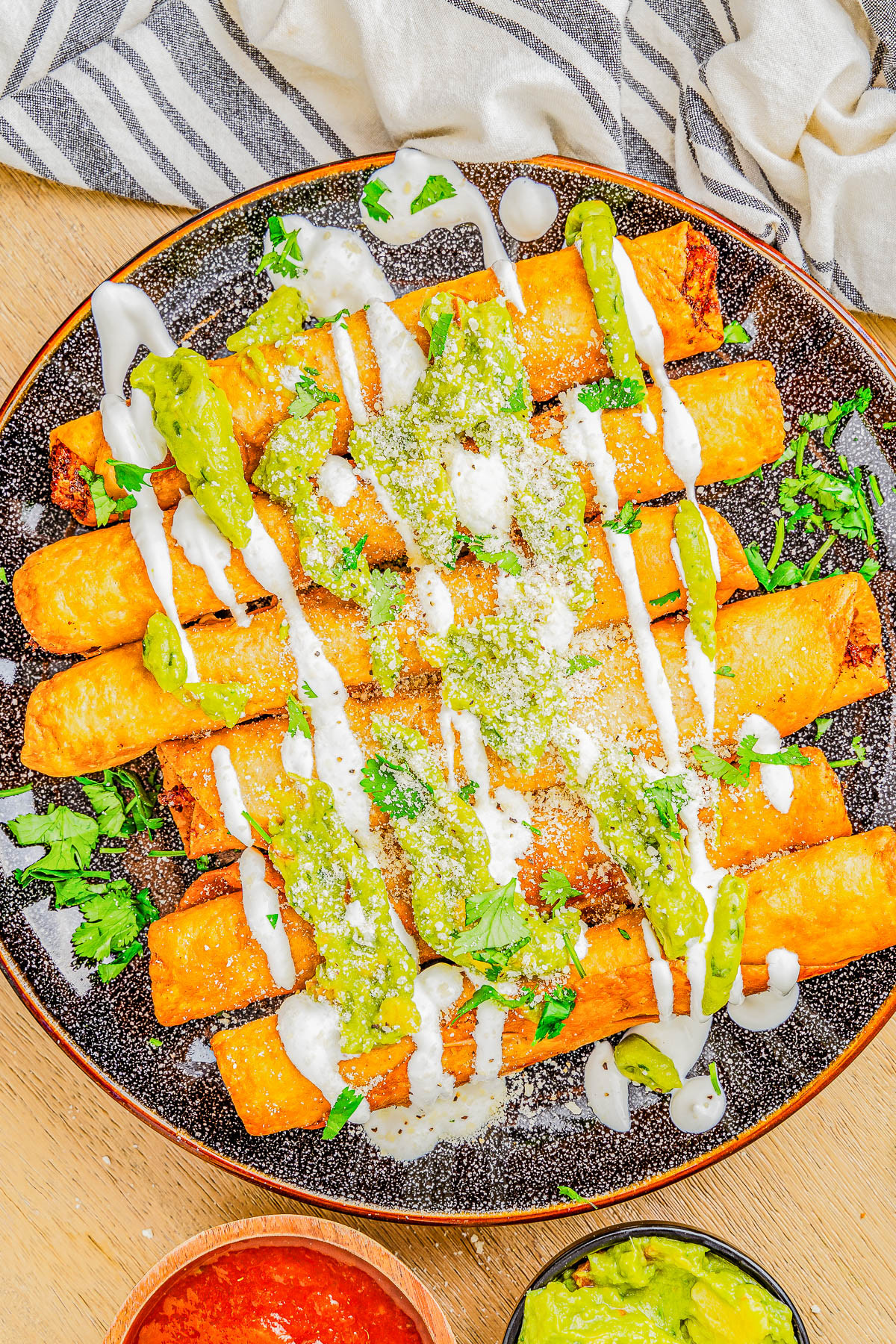 A plate of crispy taquitos topped with green sauce, white drizzle, and sprinkled cheese, garnished with chopped herbs. A bowl of red salsa and green dip is nearby.