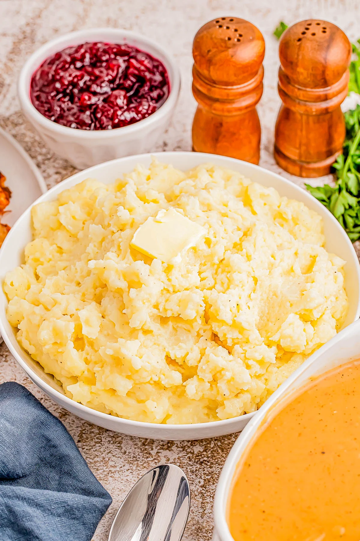 A bowl of mashed potatoes with a pat of butter, accompanied by gravy, cranberry sauce, and salt and pepper shakers on a table.