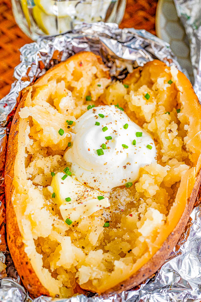 A baked potato with butter, sour cream, and chopped chives on top, wrapped in aluminum foil.