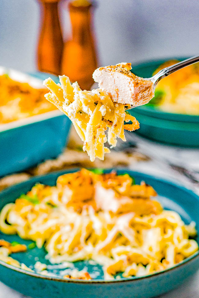 A fork holding a bite of creamy pasta with chicken, over a blue plate filled with more of the dish; two wooden pepper mills are visible in the background.