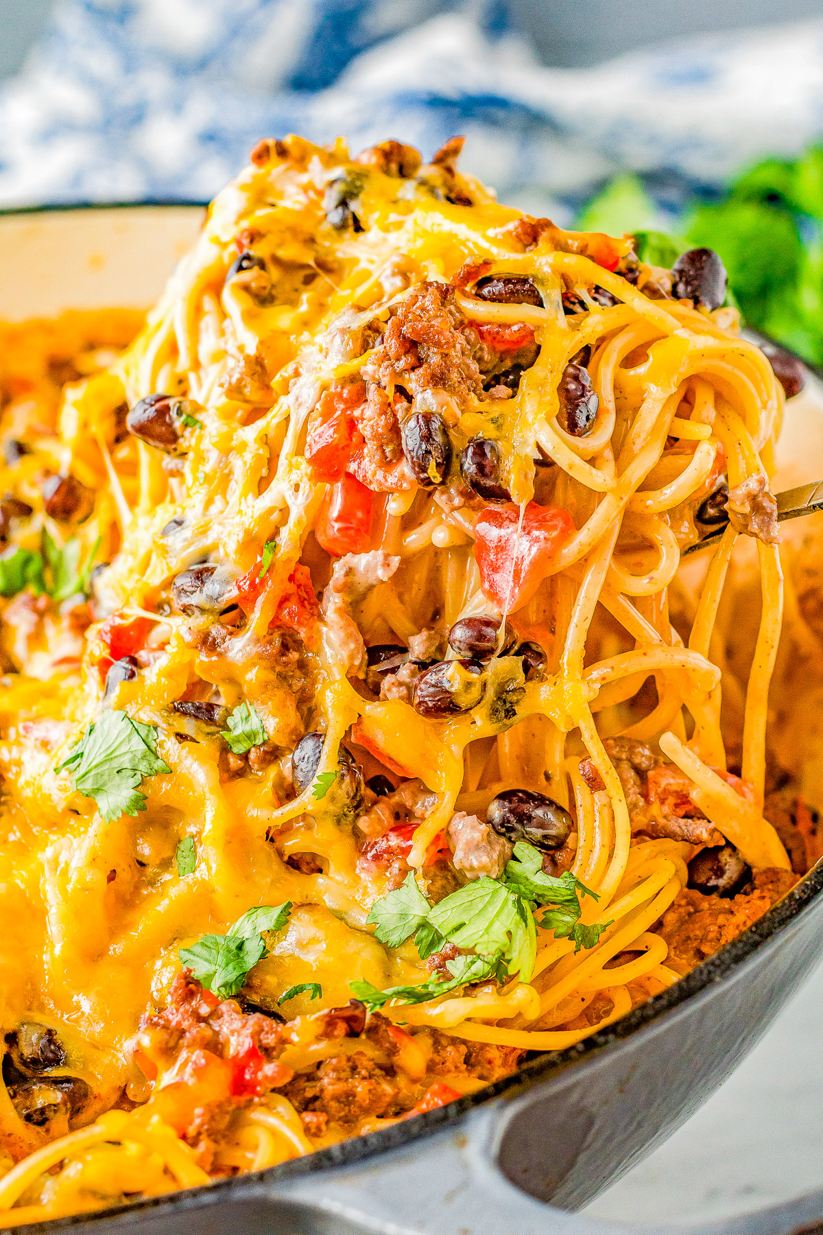 Close-up of a cheesy taco spaghetti dish with ground meat, black beans, and bell peppers, garnished with cilantro, in a skillet.