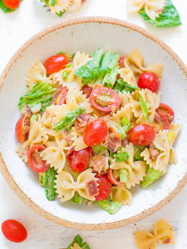 A bowl of bowtie pasta salad with cherry tomatoes, lettuce, bacon bits, and a light dressing, with additional ingredients scattered around the bowl.