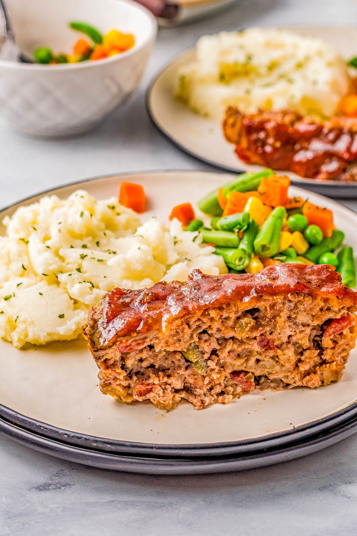 A plate with a slice of glazed meatloaf, mashed potatoes, and mixed vegetables of carrots, green beans, and peas.