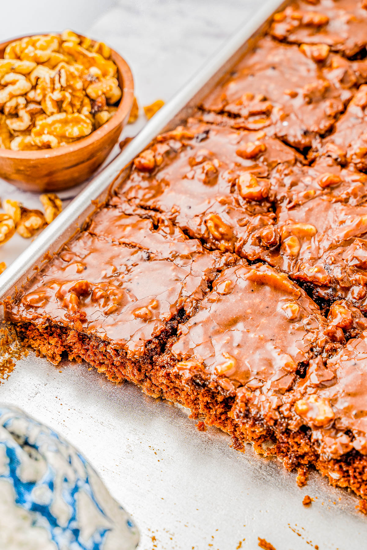 A tray of glazed chocolate brownies with walnuts, partially cut into squares. A bowl of walnuts is nearby.