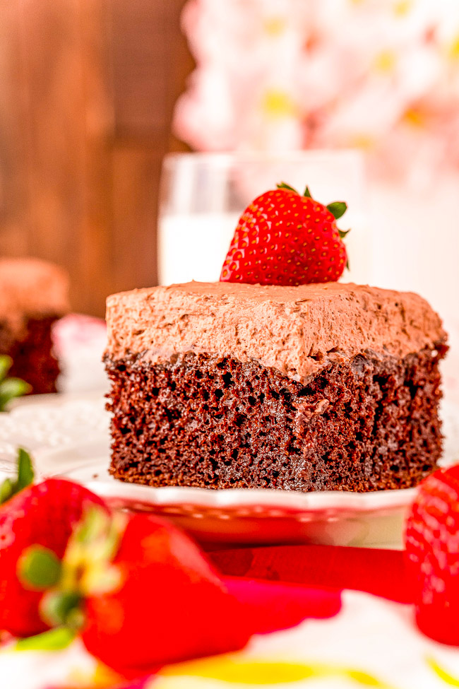 A slice of chocolate cake topped with chocolate frosting and a strawberry, with more strawberries blurred in the foreground.