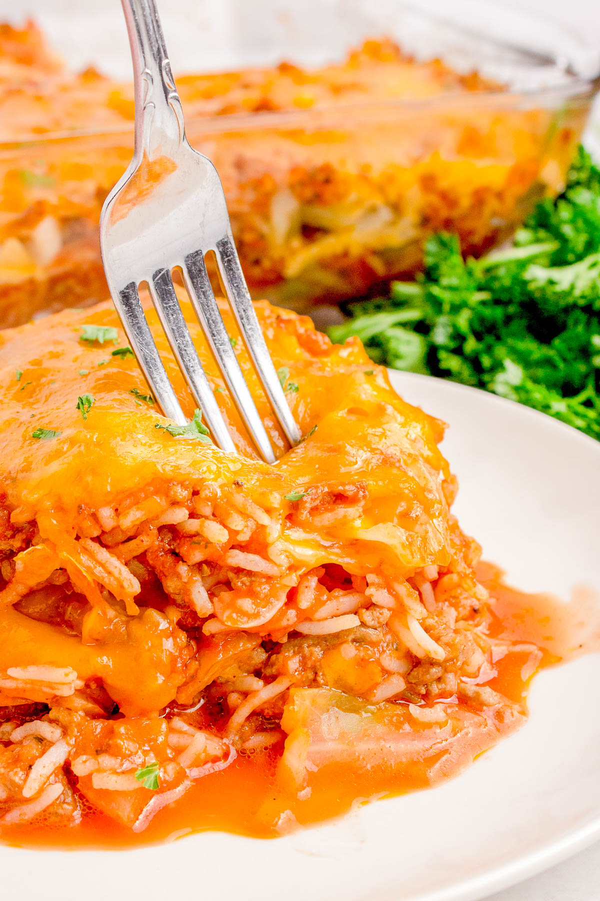 Close-up of a cheesy spaghetti casserole with a fork inserted. A side of fresh parsley is visible in the background.