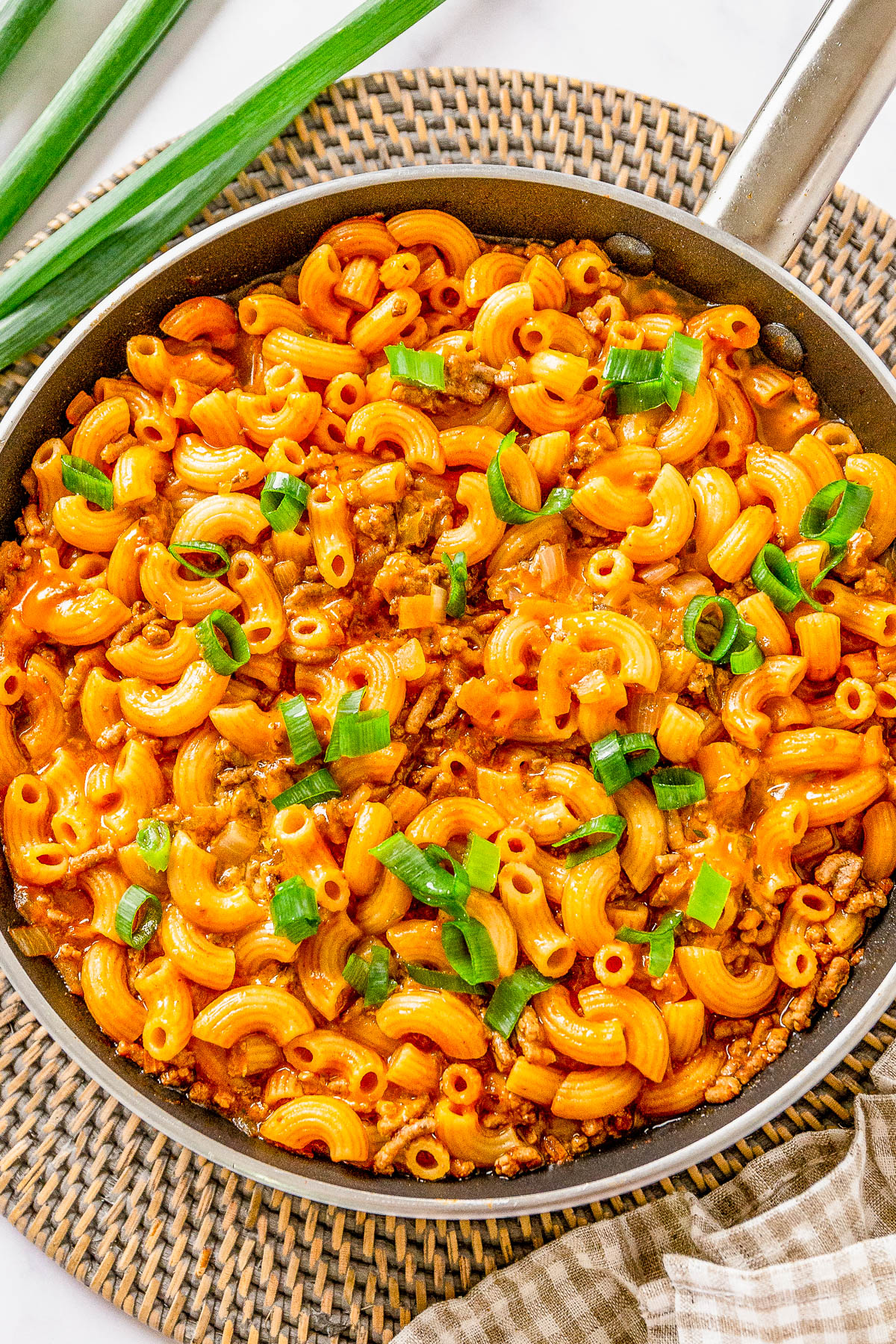 A skillet filled with cooked macaroni pasta in a creamy orange sauce, topped with chopped green onions, placed on a woven mat.