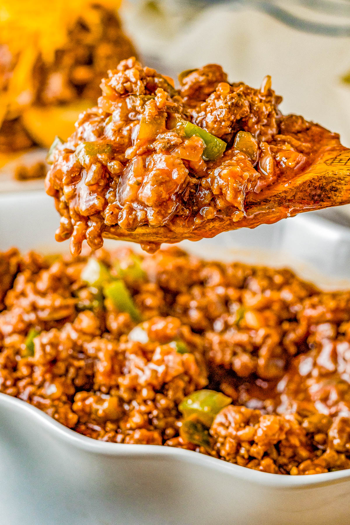 A wooden spoon holds a portion of saucy ground beef with green peppers over a dish filled with the same mixture.