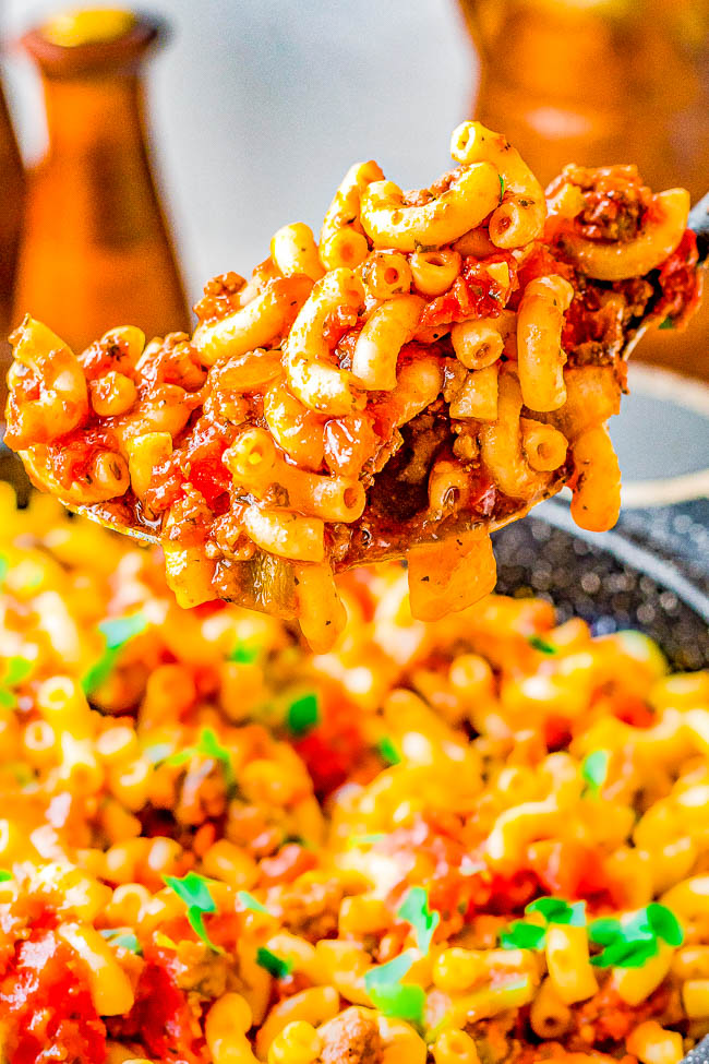 A close-up of a spoon lifting a portion of macaroni with meat sauce and herbs from a pan filled with the dish.