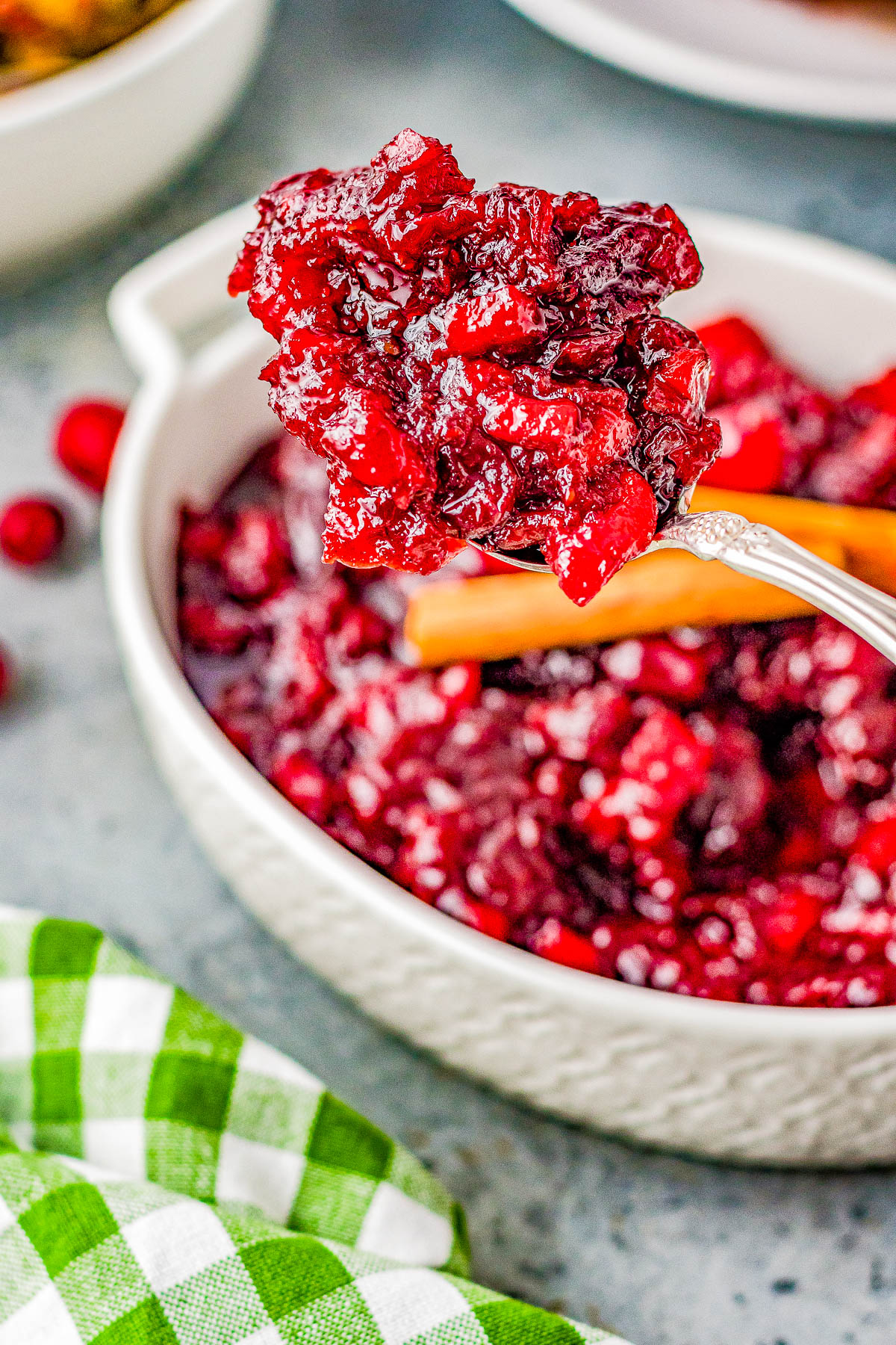 A close-up of a spoonful of cranberry sauce being held over a bowl filled with the sauce, with a green and white checkered cloth in the foreground.