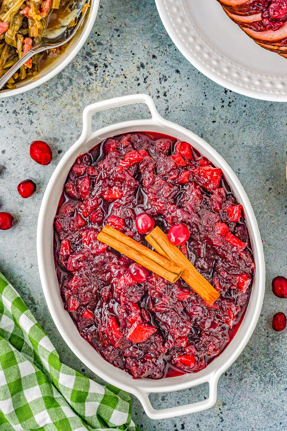 A dish of cranberry sauce garnished with cinnamon sticks, surrounded by other dishes on a grey surface with a green checked cloth nearby.