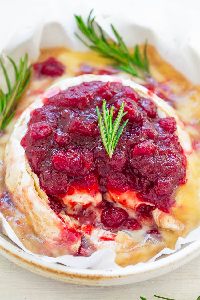 Close-up of baked brie topped with cranberry sauce and garnished with a sprig of rosemary, served in a white dish.