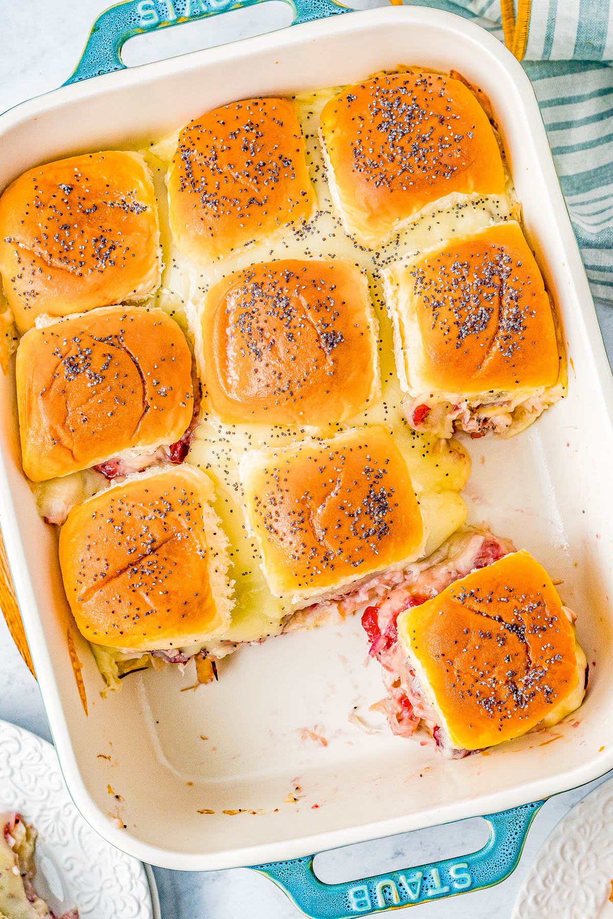 A baking dish with nine baked slider sandwiches topped with poppy seeds. The sandwiches are partially sliced, revealing cheese and meat inside.