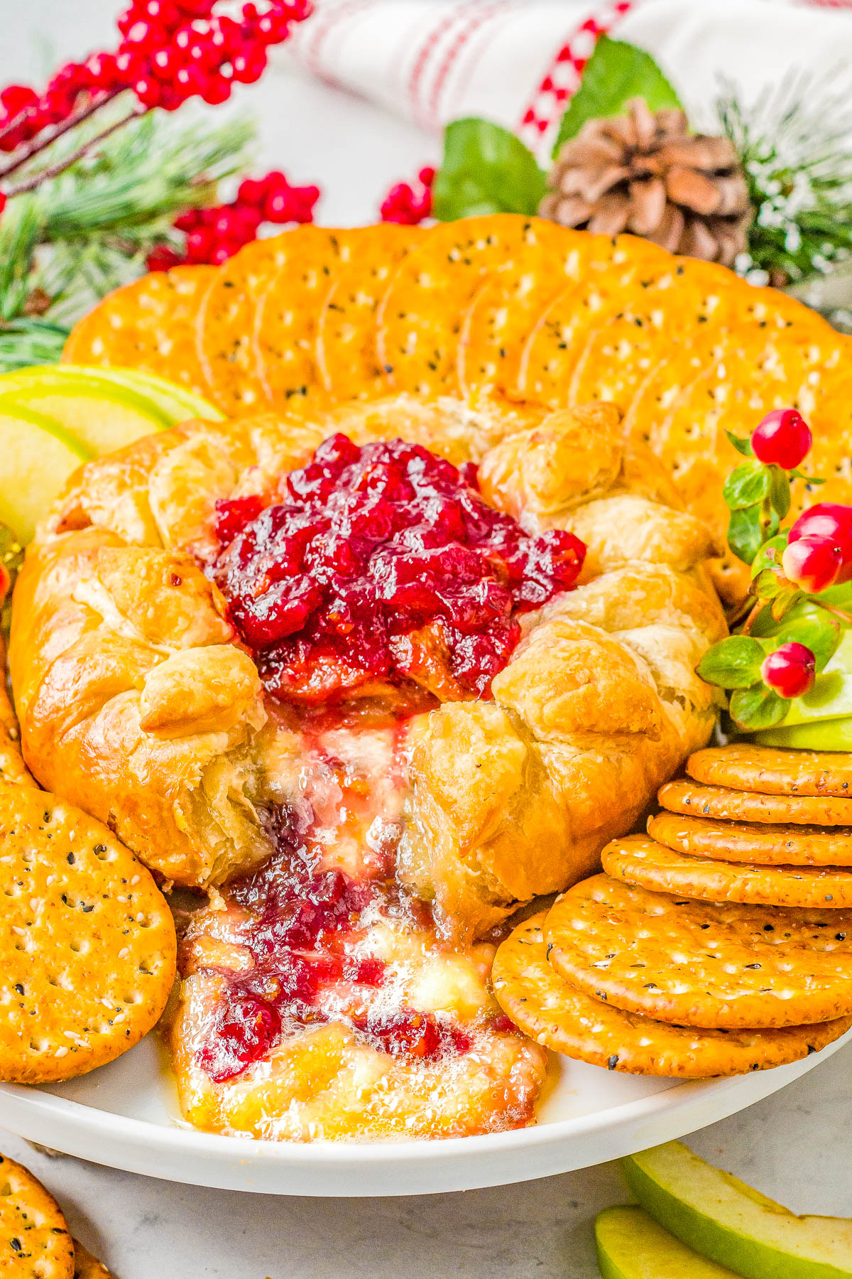 A plate with baked brie topped with cranberry sauce, surrounded by round crackers, green apple slices, and holiday decorations.