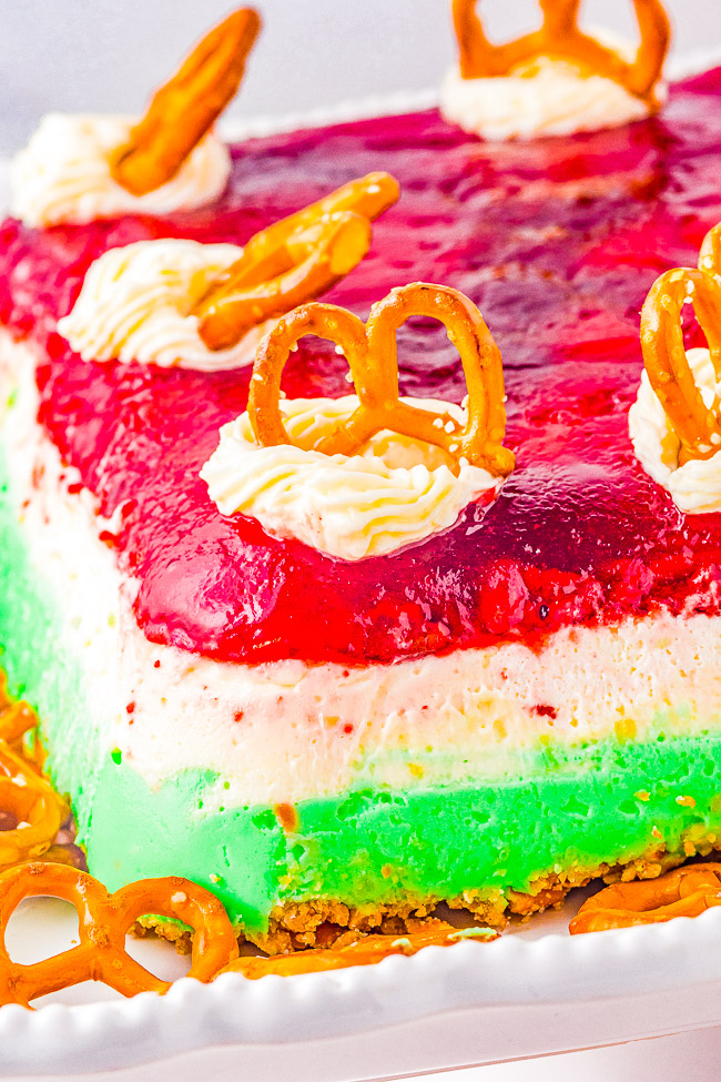 A colorful layered dessert with green, white, and red layers, topped with dollops of cream and pretzels, presented on a white tray.