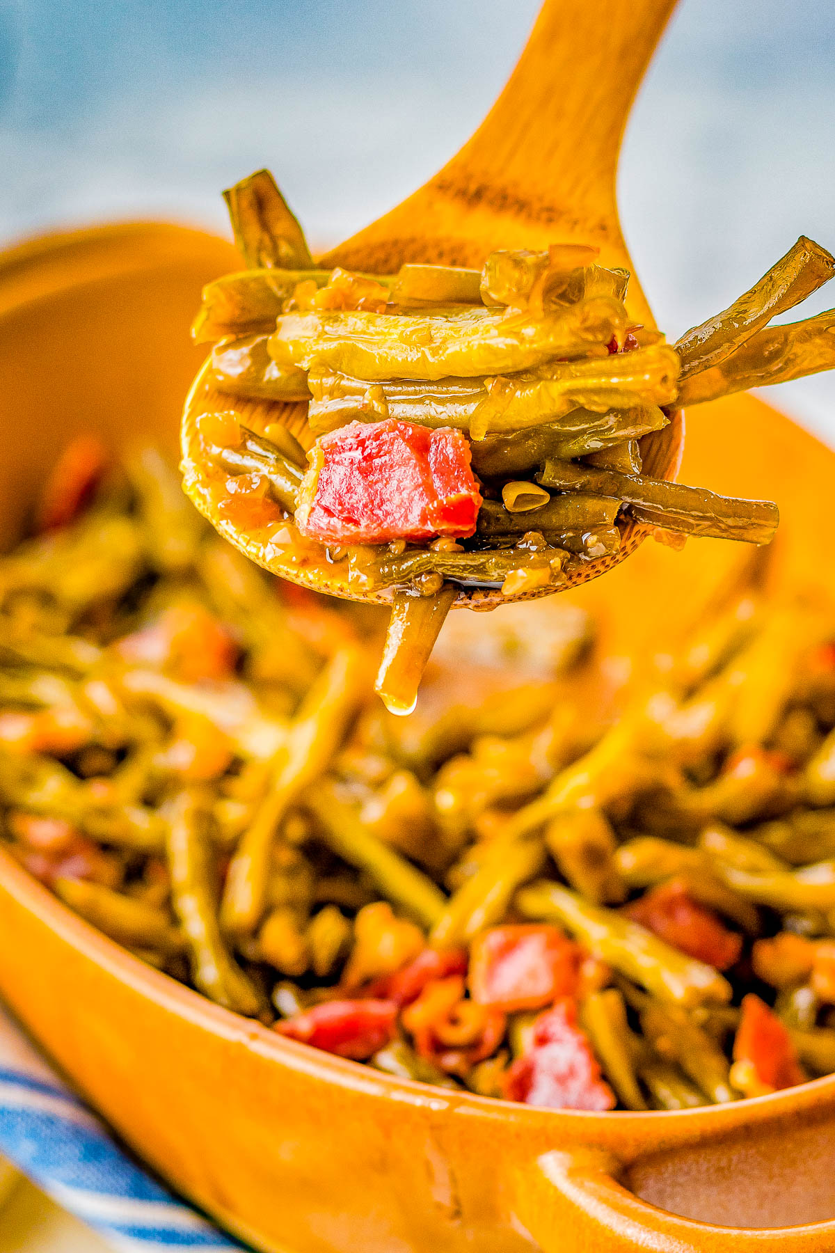 A wooden spoon lifting a portion of cooked green beans with diced tomatoes from a dish.