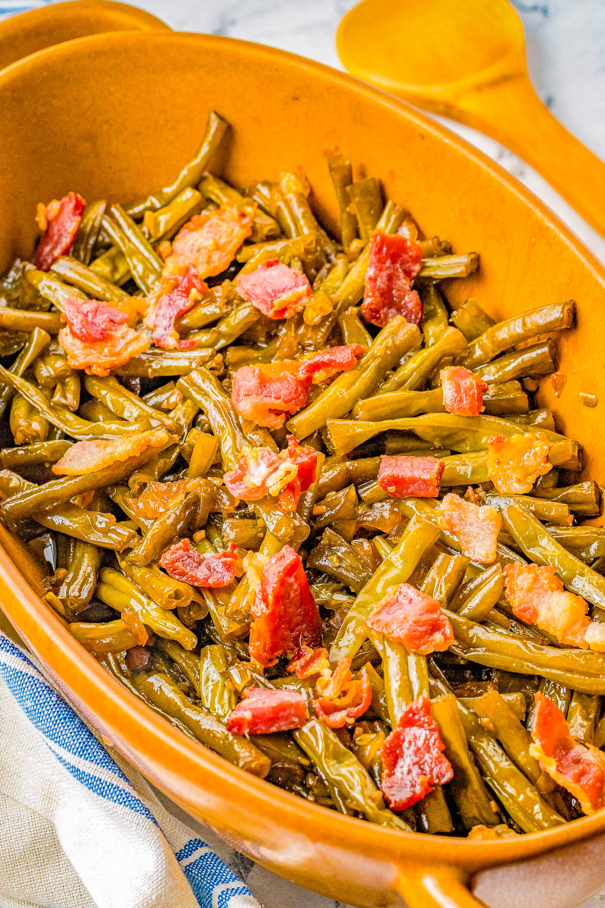 A ceramic dish filled with cooked green beans and chunks of bacon, accompanied by a wooden spoon in the background.