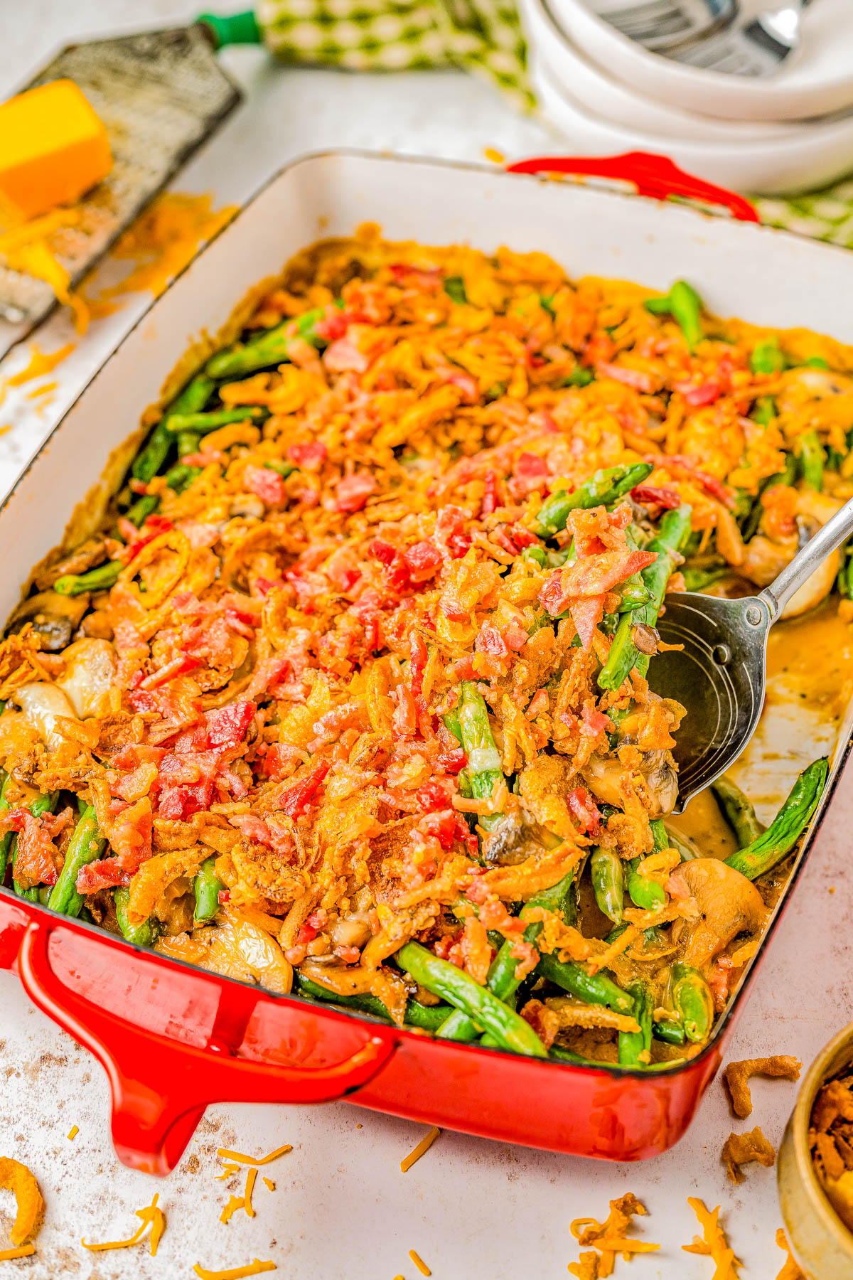 A close-up of a red casserole dish filled with green bean casserole, topped with crispy fried onions and bits of bacon. A spoon is partially inserted into the dish, about to serve a portion.