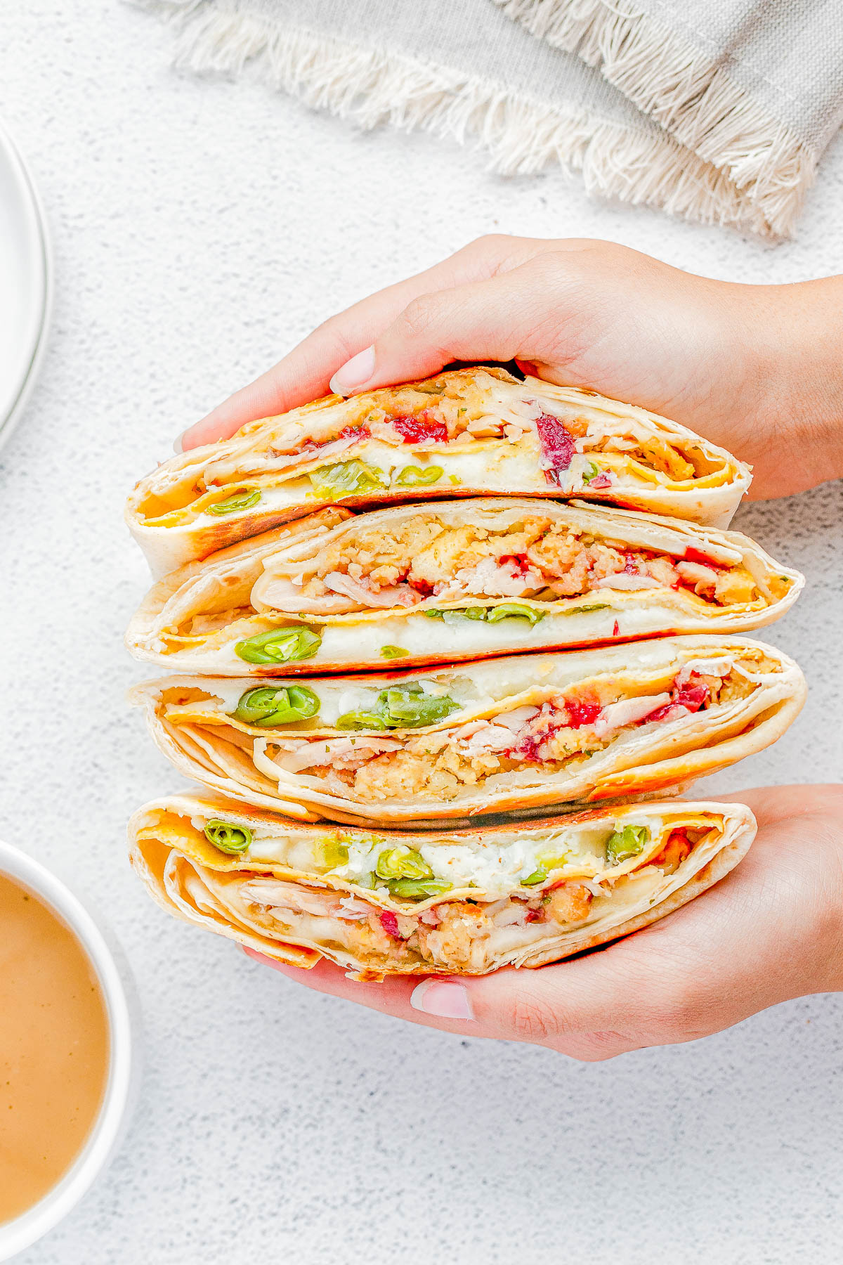 Hands holding three folded tortillas filled with mixed ingredients, including green peppers and red bits, displayed on a light surface with a bowl of sauce nearby.