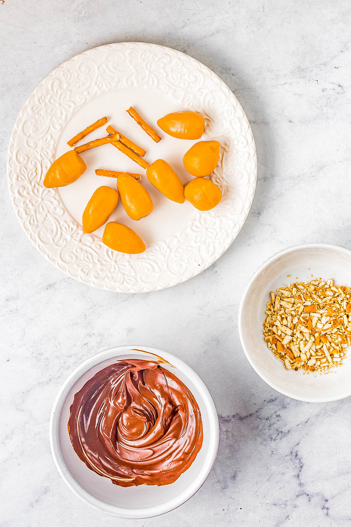 A plate with small orange bite-sized snacks and pretzel sticks, a bowl of crushed pretzels, and a bowl of melted chocolate on a marble surface.