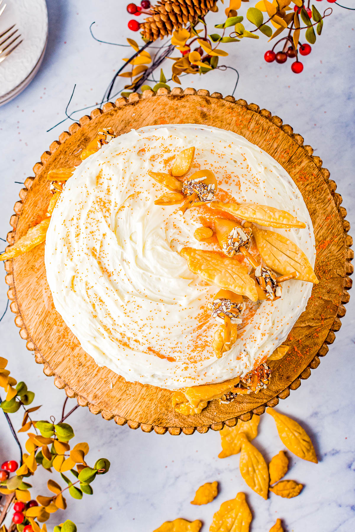 A frosted cake topped with orange decorations and sprinkled with nut pieces is displayed on a round wooden serving board. Decorative autumn elements surround the cake.