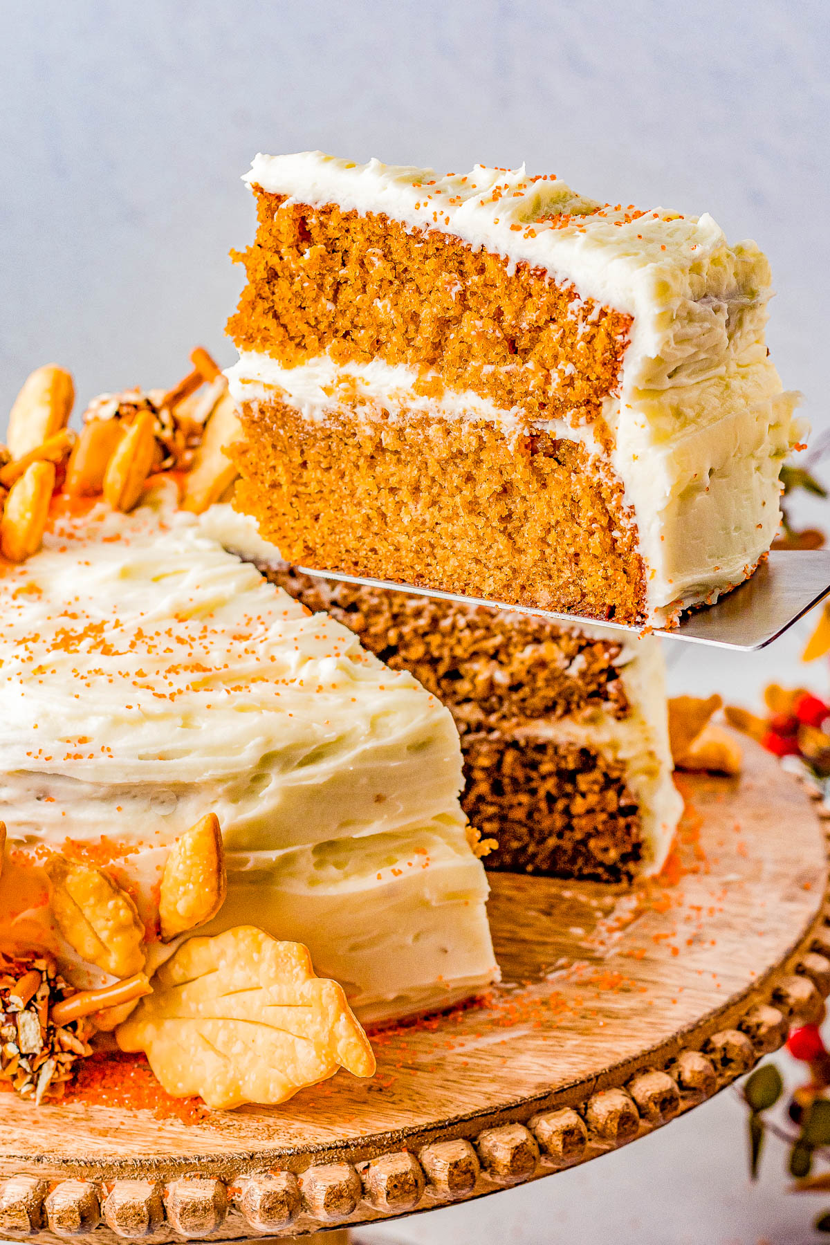 A slice of a two-layered carrot cake with cream cheese frosting is being lifted from the whole cake, which is decorated with nuts and orange sprinkles.