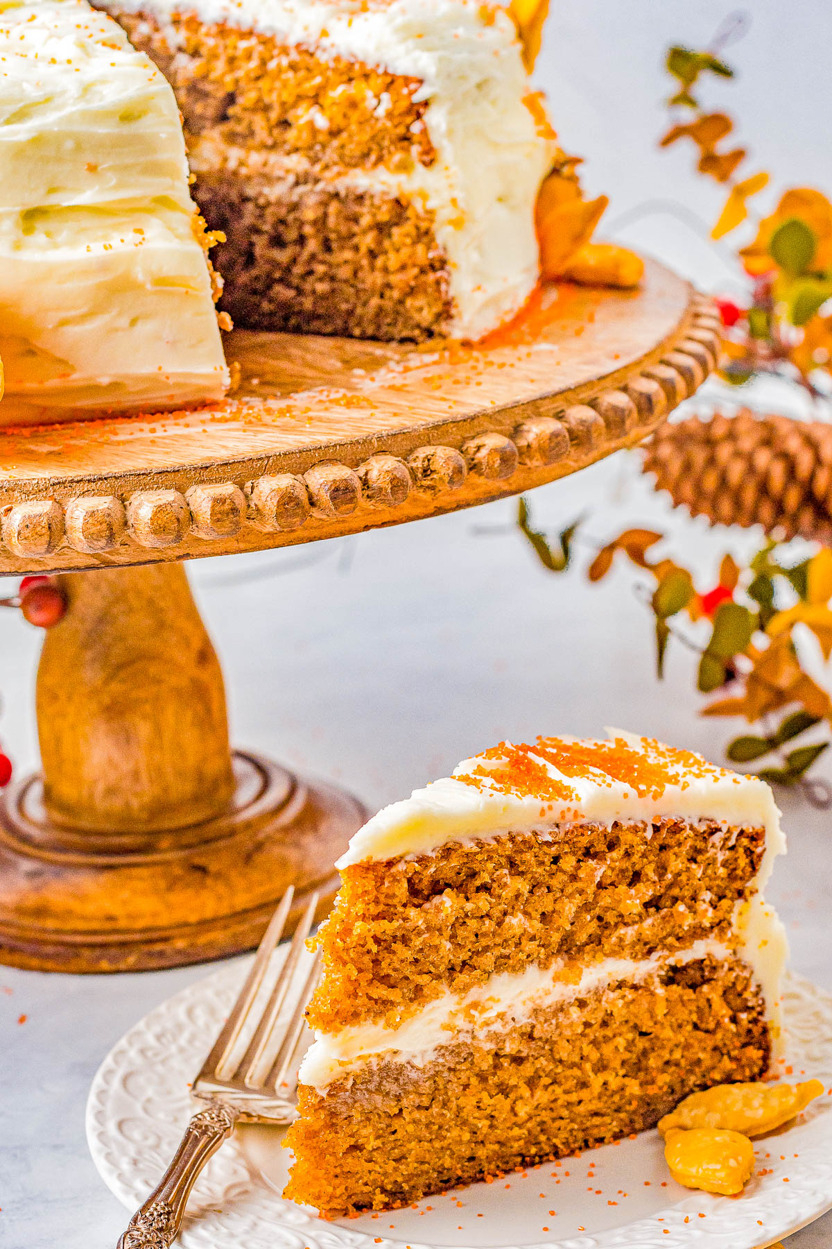 A slice of frosted layer cake is served on a white plate with a fork, in front of a wooden cake stand holding the remaining cake. The background has autumn-themed decor.