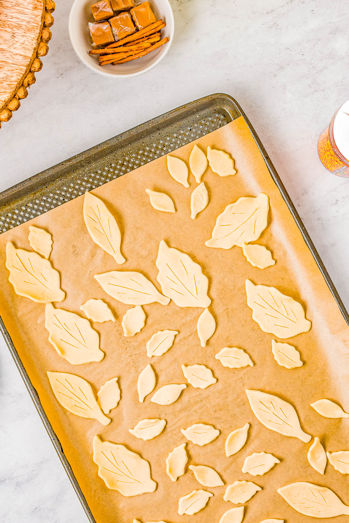 A baking tray with leaf-shaped pastry cutouts on parchment paper next to a bowl of caramel candies.