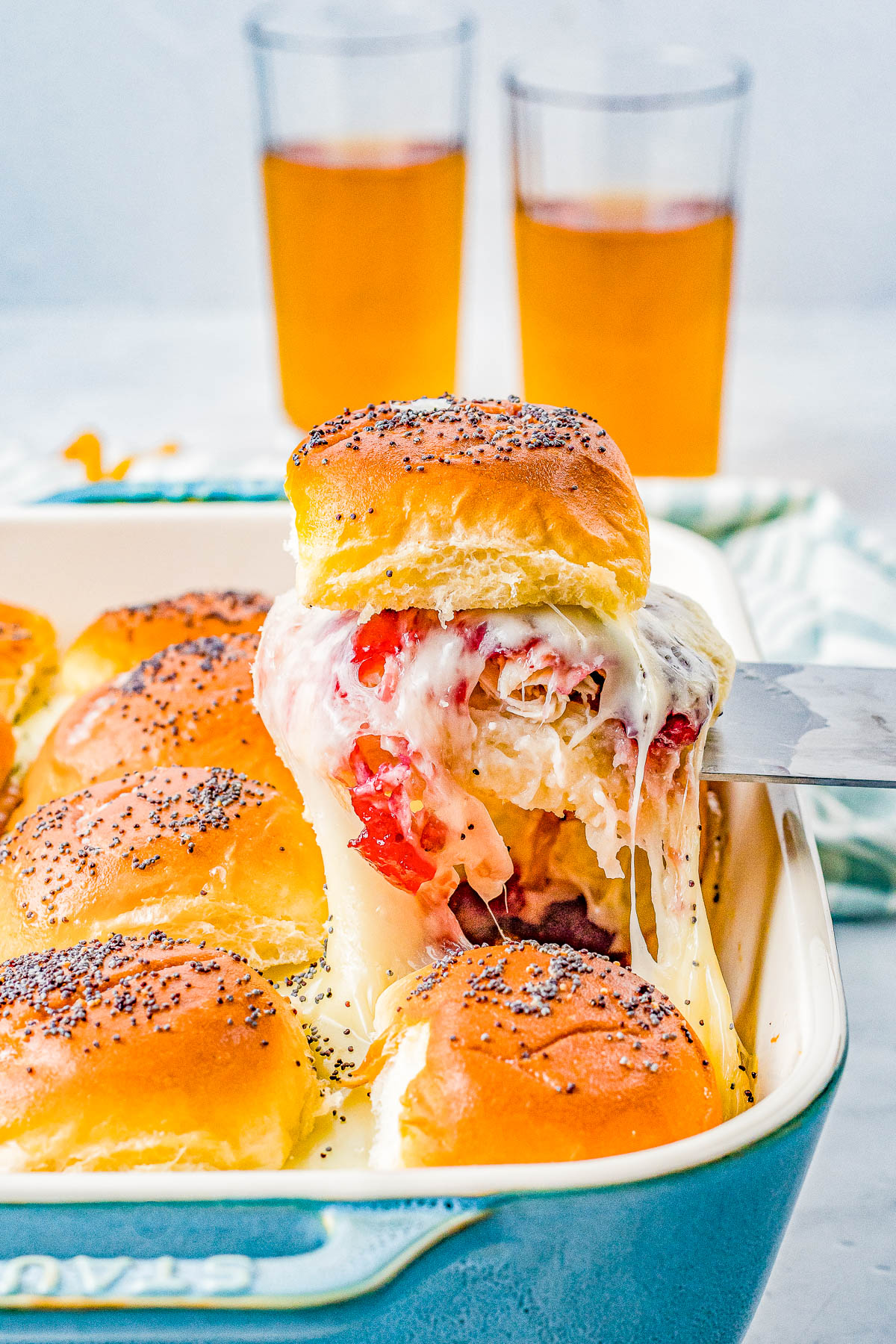 A pan of baked sliders topped with poppy seeds, featuring melted cheese and tomato slices, with two glasses of amber-colored beverages in the background.