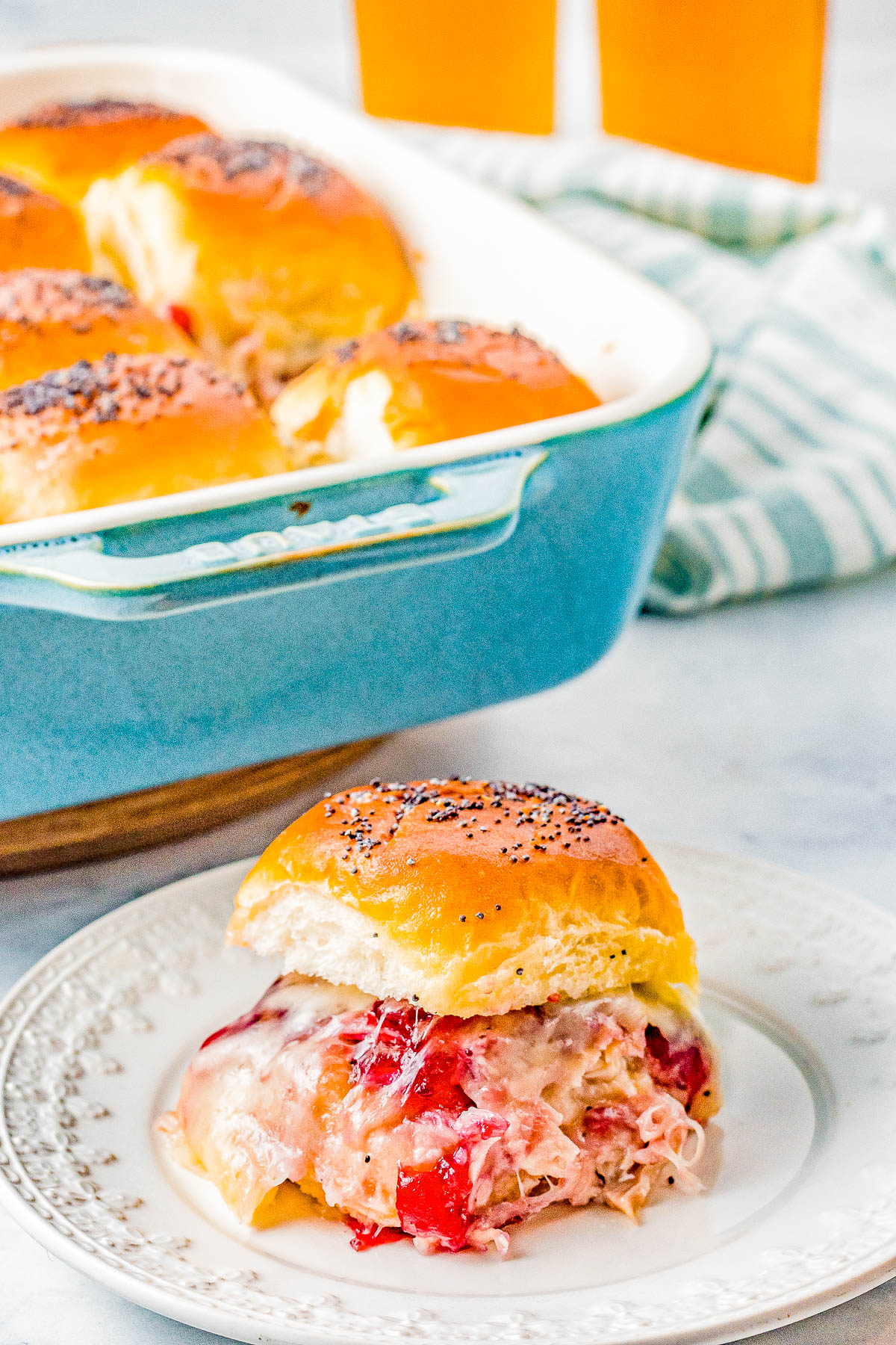 A sandwich filled with cheese and ham topped with poppy seeds on a plate. A blue baking dish with more sandwiches is in the background, along with glasses of orange liquid.