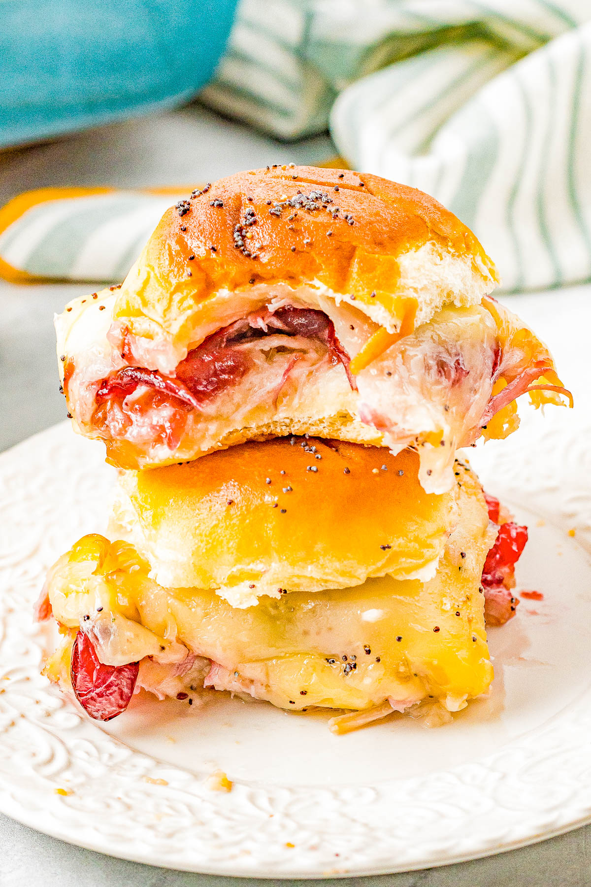 Two stacked ham and cheese sliders on a white plate, topped with poppy seeds and leaking cheese.