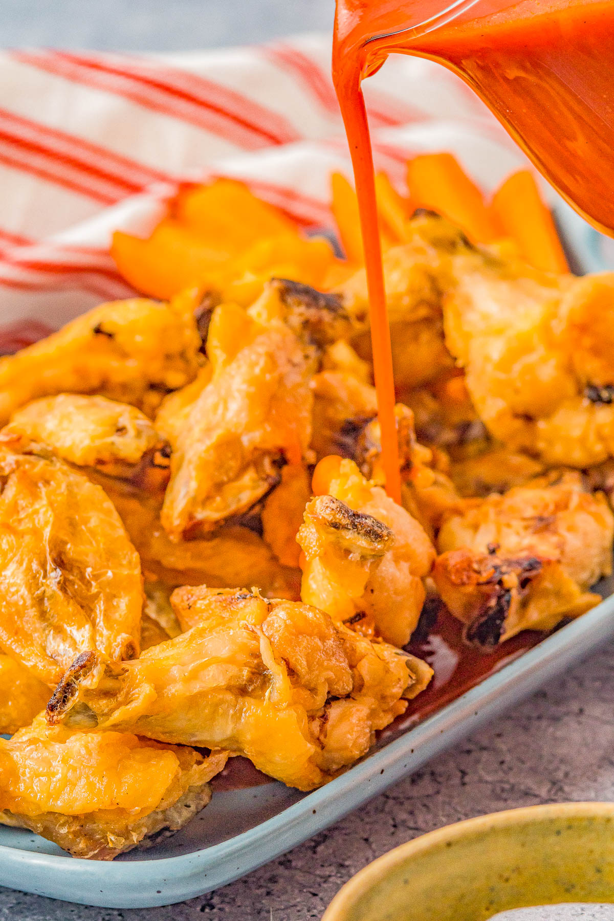 Hot sauce being poured over crispy chicken wings on a plate with carrot sticks in the background.